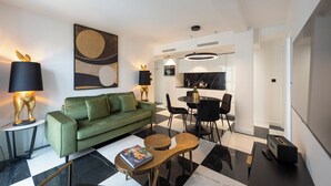 Sala de estar moderna com um sofá verde, uma mesa de centro e um espaço de jantar com uma mesa redonda e cadeiras. Há uma grande pintura abstrata na parede e um abajur com design de coelho em uma mesinha lateral.