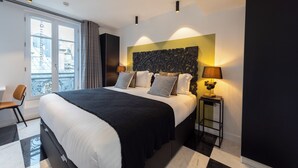 Habitación de hotel moderna con una cama grande, una mesita de noche con lámpara y vistas a la ciudad a través de un ventanal amplio.