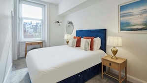 Quarto de hotel com uma cama grande, duas mesas de cabeceira com abajures, vista para a cidade pela janela e um quadro na parede.