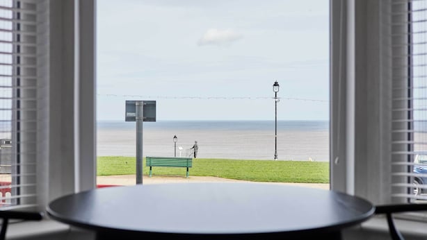 Vista da janela com uma mesa em primeiro plano, um banco e uma área gramada ao fundo, onde se vê um poste de luz e uma estátua.
