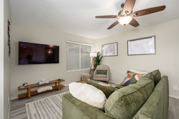 A living room with a green sofa, a beige armchair, a wooden bench, a TV, and a ceiling fan. - Nicely appointed condo on the golf course and running/biking path! (Scottsdale)