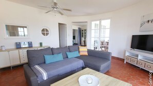 A living room with a grey sofa, a wooden coffee table, a TV stand, and a ceiling fan. - On the beach 2 bedrooms flat 4/6 persons (PORT GRIMAUD)