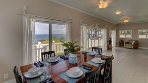 A dining area with a table set for four, large windows with curtains, a ceiling fan, and a view of the sea. - Paradise Ocean View, 2Br/2Ba (Keaau)