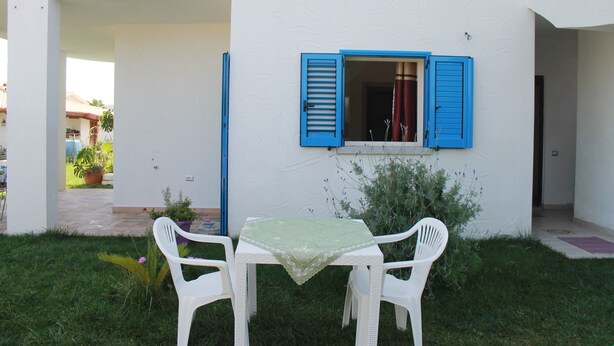 Zona d'estar a l'aire lliure amb una taula i dues cadires, una finestra amb persianes blaves i una paret blanca.