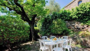 A stone wall with a rustic building in the background, a tree in the foreground, and a white table with chairs set up outdoors. - Charming House In Garde Freinet With Garden (La Garde-Freinet)