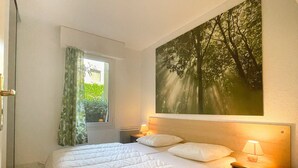 A hotel room with a large bed, a nightstand with a lamp, a window with curtains, and a painting of trees on the wall.