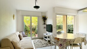 A living room with a sofa, a dining table with chairs, a TV, and a view of greenery through the windows.