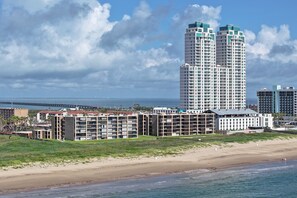 A coastal hotel with multiple high-rise buildings, a beach, and a bridge in the background. - Sea Vista II #224 | Cozy Beachfront Condo • Direct Beach Access • Two Community Pools (South Padre Island)
