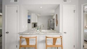 A compact kitchen with a marble countertop, two wooden bar stools, a refrigerator, and a coffee maker. - Villa Stone Creek 2 Bedroom Condo by RedAwning (Scottsdale)