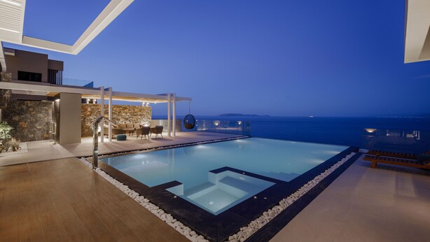 A modern infinity pool with a clear view of the ocean, a stone wall, and a covered seating area with a hanging chair. - Villa Nereid 5 bedroom villa with infinity heated pool (Rodia)