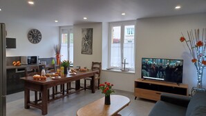 Ein modernes Wohnzimmer mit Esstisch aus Holz, einer Couch, einem Fernseher und einer Küchenzeile mit Mikrowelle und Obstschale.