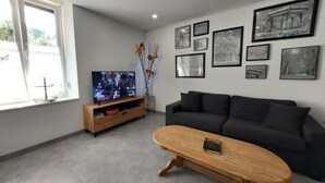 Ein modernes Wohnzimmer mit einem dunkelgrauen Sofa, einem Holztisch und einem Fernseher auf einem hölzernen Schrank. An den Wänden befinden sich gerahmte Bilder und auf einem kleinen Tisch steht eine Vase mit Blumen.
