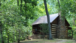 Une cabane rustique en rondins de bois, posée sur des fondations en pierre, entourée d’une zone boisée et accessible par un sentier en pierre.