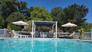 Une piscine d’un bleu éclatant, bordée de chaises longues et d’ombrelles, et entourée d’une clôture blanche et d’arbres.