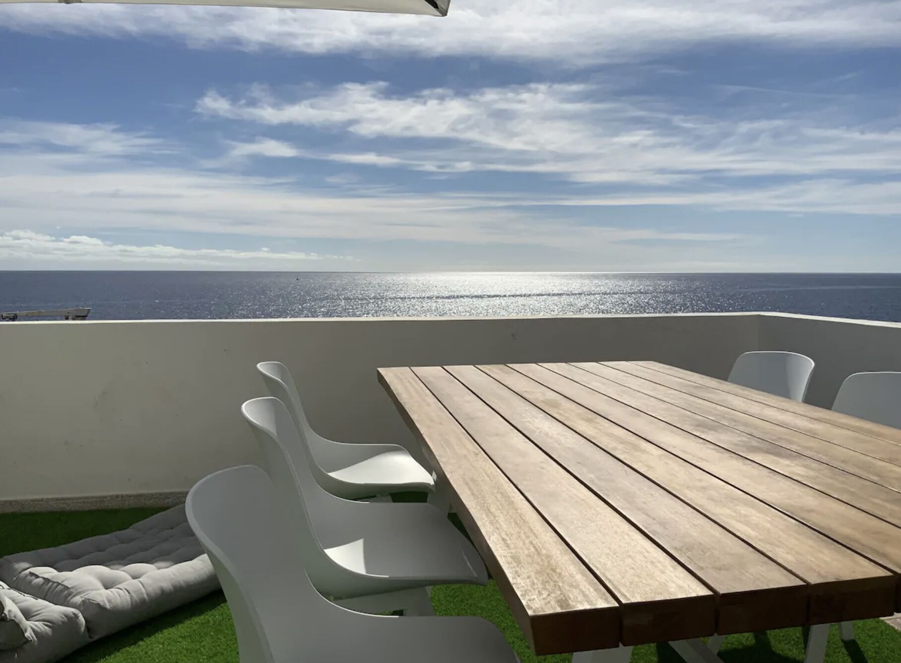 Une table en bois, entourée de chaises blanches, sur un balcon donnant sur la mer.