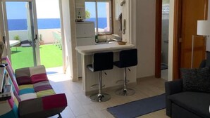 Una sala de estar moderna con un sofá, una mesa de centro y un televisor. Hay una cocina con un refrigerador, un fregadero y una encimera. La habitación tiene un ventanal con vista al océano.