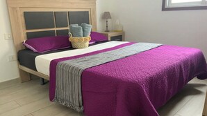 Una cama bien hecha con un edredón morado y una manta gris, un cabecero de madera y una mesita de noche con una lámpara y una caja de pañuelos.