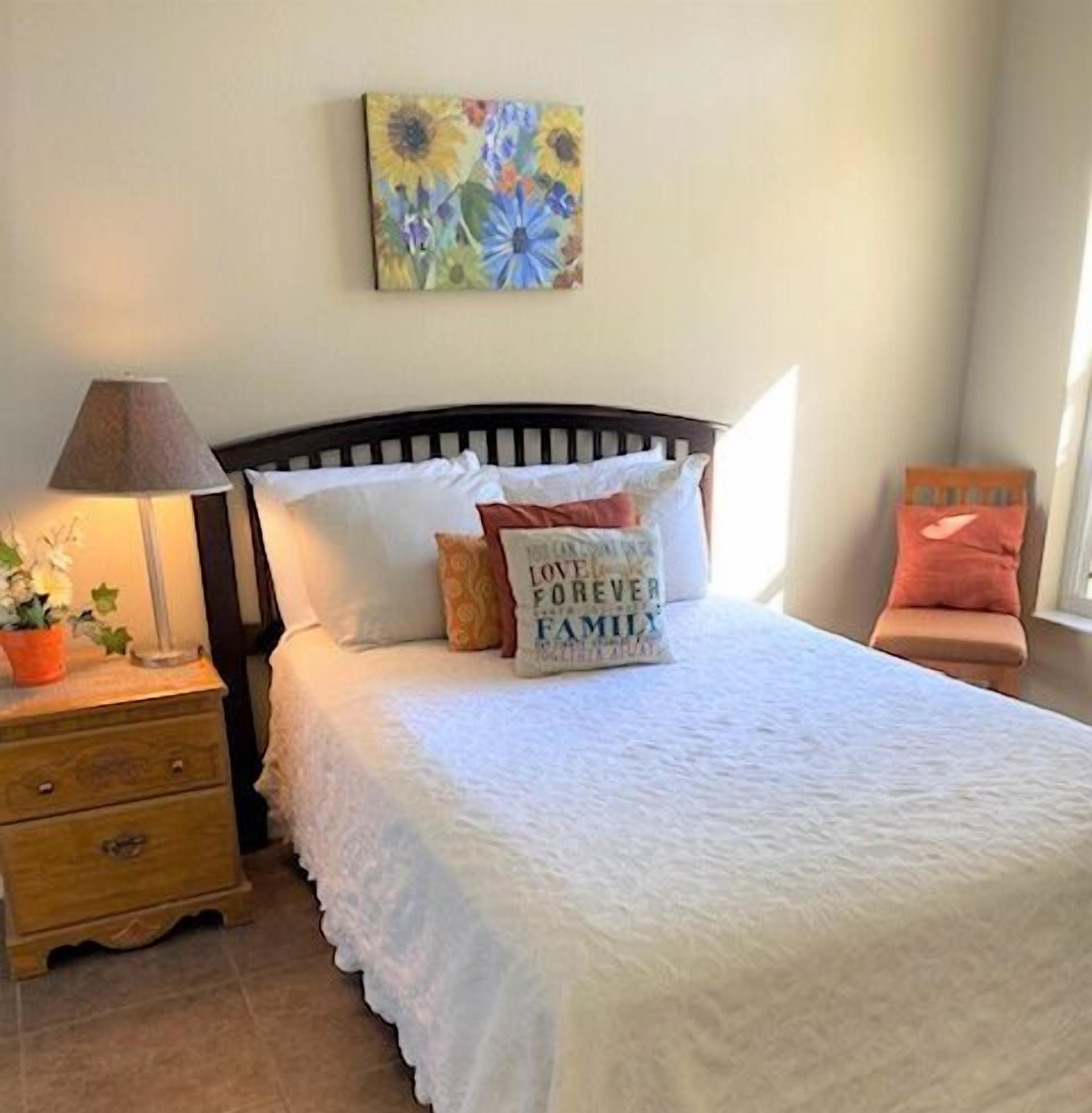 A bedroom with a bed, pillows, a nightstand, a lamp, and a painting on the wall.