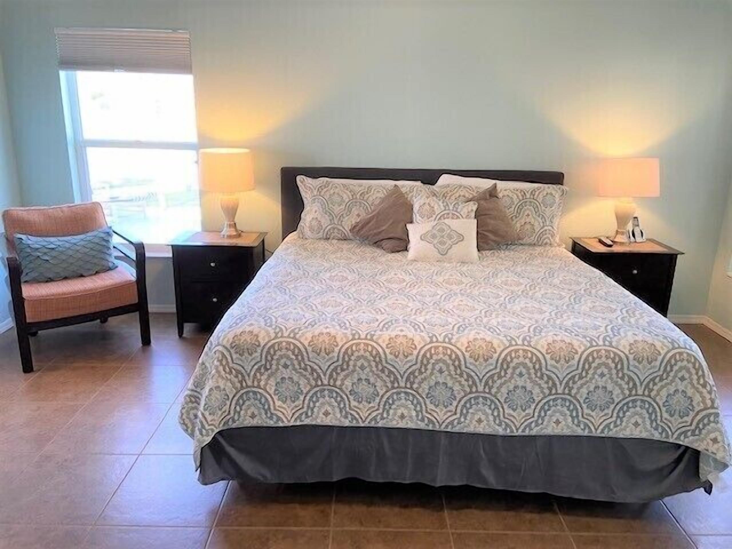 A bedroom with a large bed, two bedside tables, a chair, and a window with blinds.