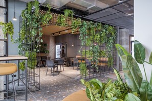 An indoor space with a modern design, featuring a brick wall, metal grid structures with greenery, and a mix of seating arrangements. - Citybox Antwerp City Center (Antwerp)