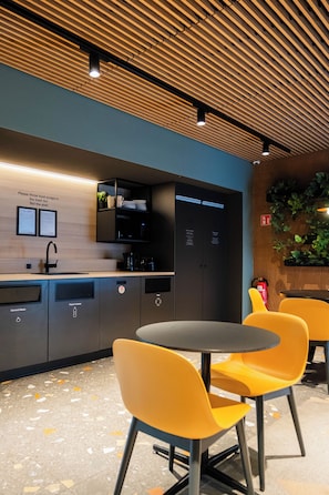 A modern kitchen with a wooden ceiling, a sink, and a coffee machine. There are two yellow chairs and a table.