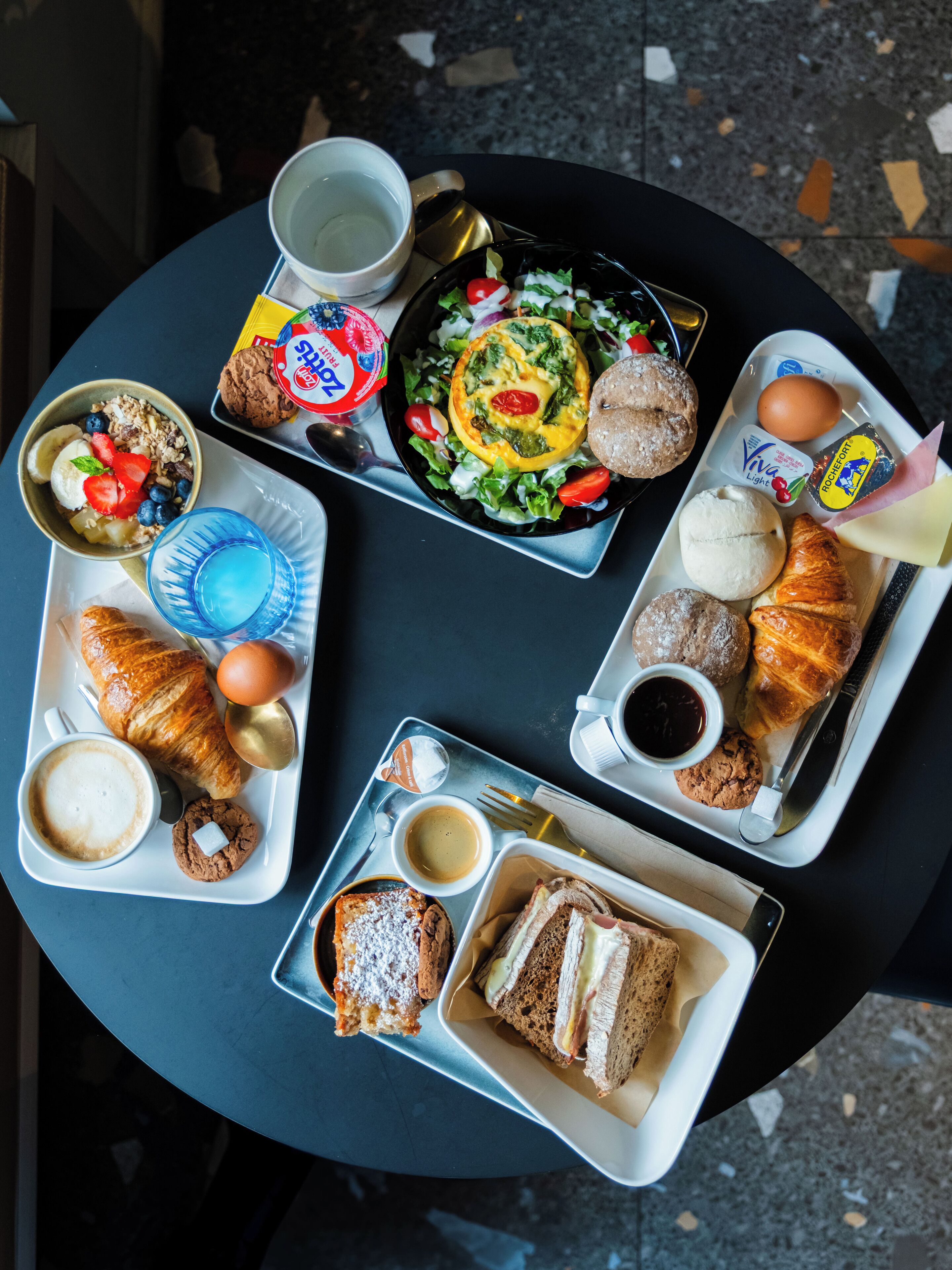 Una mesa con diversos alimentos para el desayuno, incluyendo cruasanes, un sándwich, un bol de ensalada y un plato con un cruasán y una galleta.