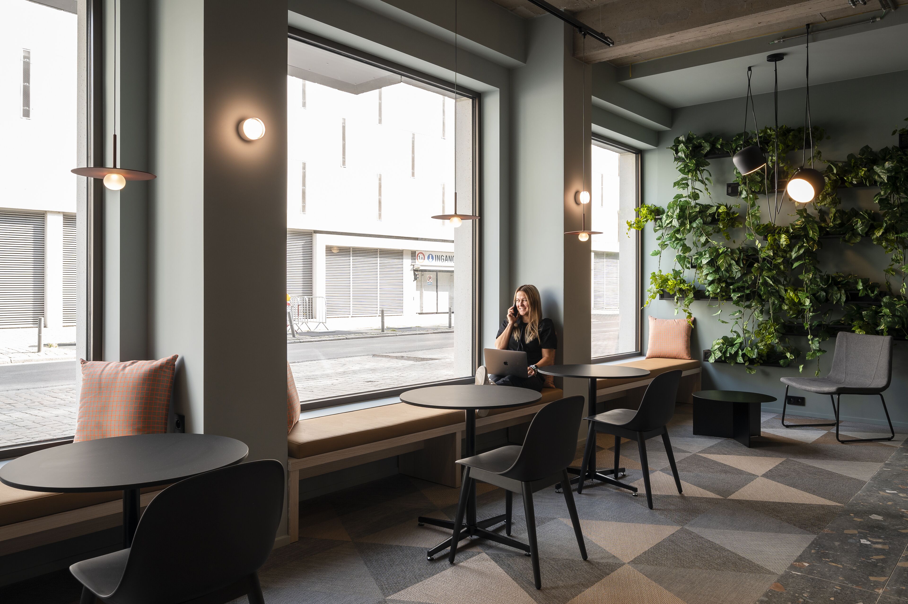 A modern cafe with round tables, comfortable seating, large windows, and indoor plants.