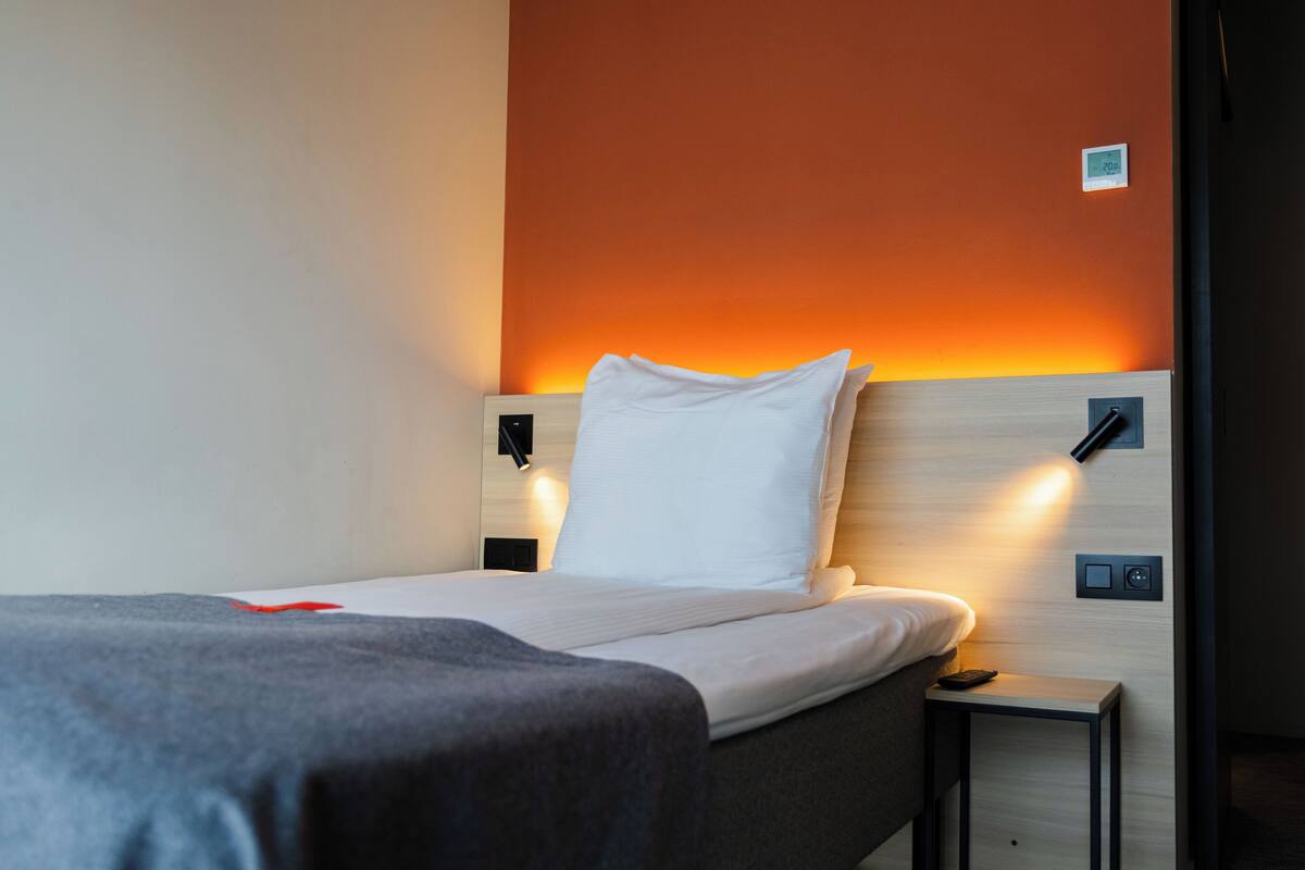 Een net opgemaakt bed met een wit kussen en een grijze deken, een nachtkastje met een afstandsbediening, en een wandlamp.