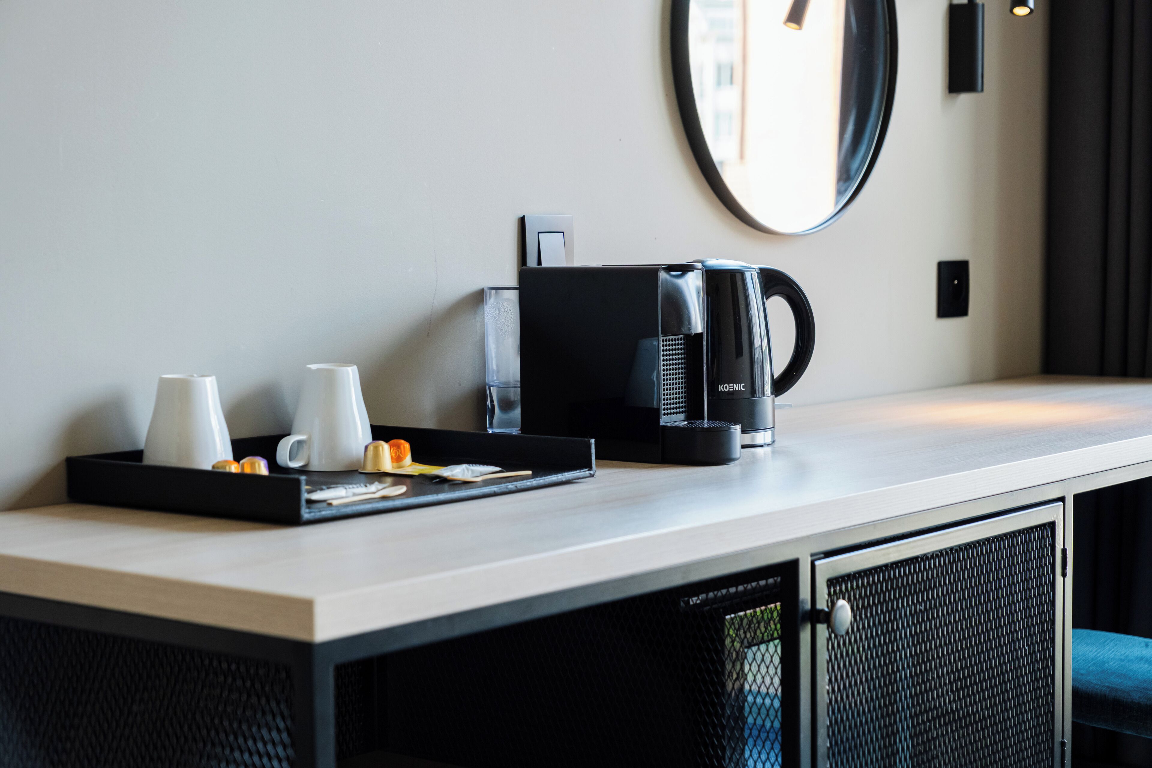 A hotel room with a kitchenette area, including a coffee maker, a tray with cups and a small orange, and a mirror on the wall.