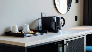 A hotel room with a kitchenette area, including a coffee maker, a tray with cups and a small orange, and a mirror on the wall.