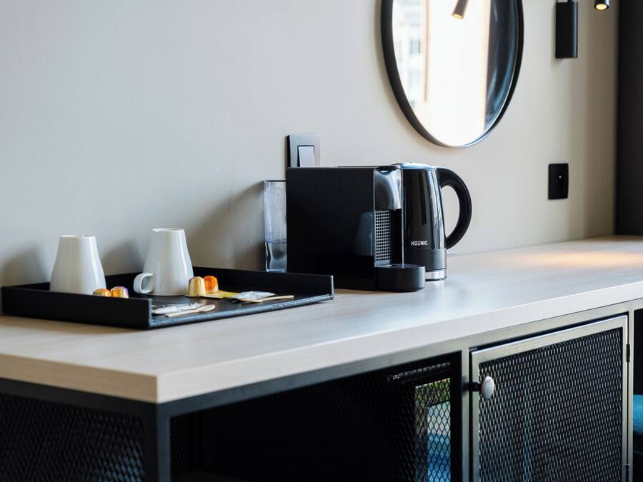 A hotel room with a kitchenette area, including a coffee maker, a tray with cups and a small orange, and a mirror on the wall.