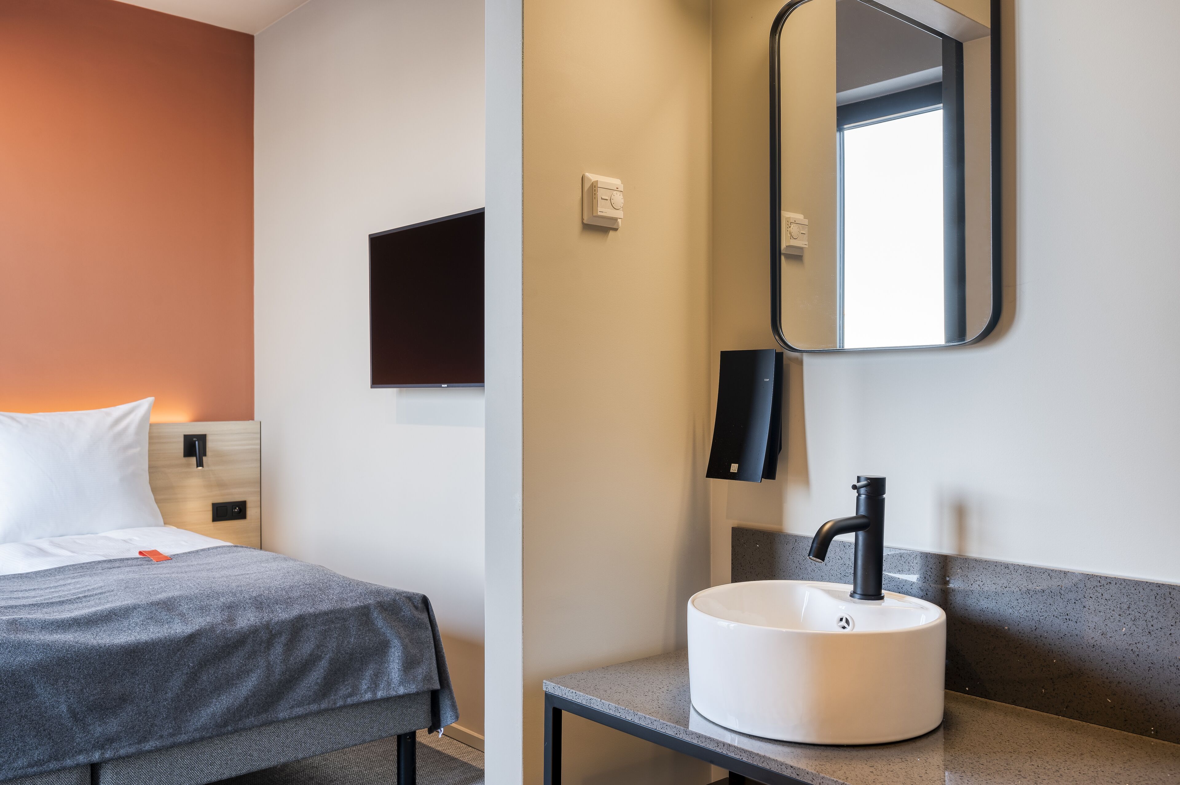 Une chambre d’hôtel comprenant un lit, une table de chevet, un lavabo moderne, un miroir et une lampe fixée au mur.