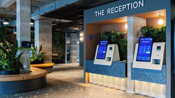 Een receptie met een bord waarop 'THE RECEPTION' staat, twee self-service kiosken en binnenshuis planten.