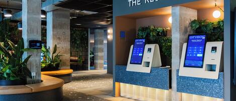 A reception area with a sign reading "THE RECEPTION," two self-service kiosks, and indoor plants.