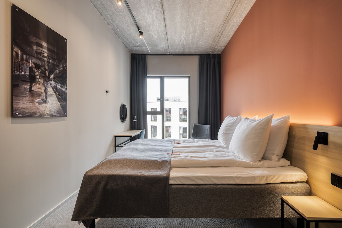 Een moderne hotelkamer met een groot bed, een bureau en uitzicht op de stad door een groot raam.