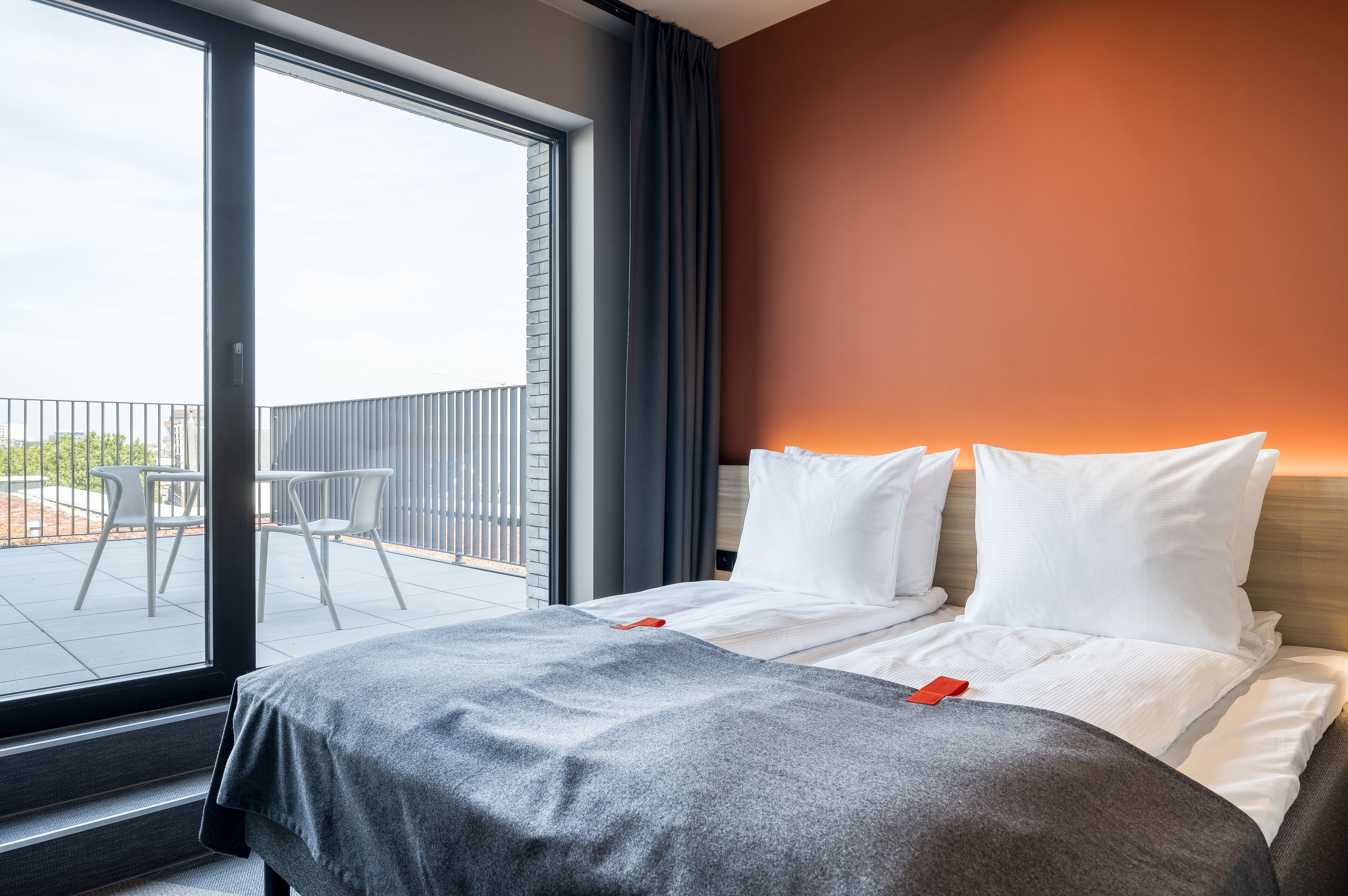 Ein Hotelzimmer mit großem Fenster, einem Balkon mit Stühlen sowie einem Bett mit weißen Kissen und einer grauen Decke.