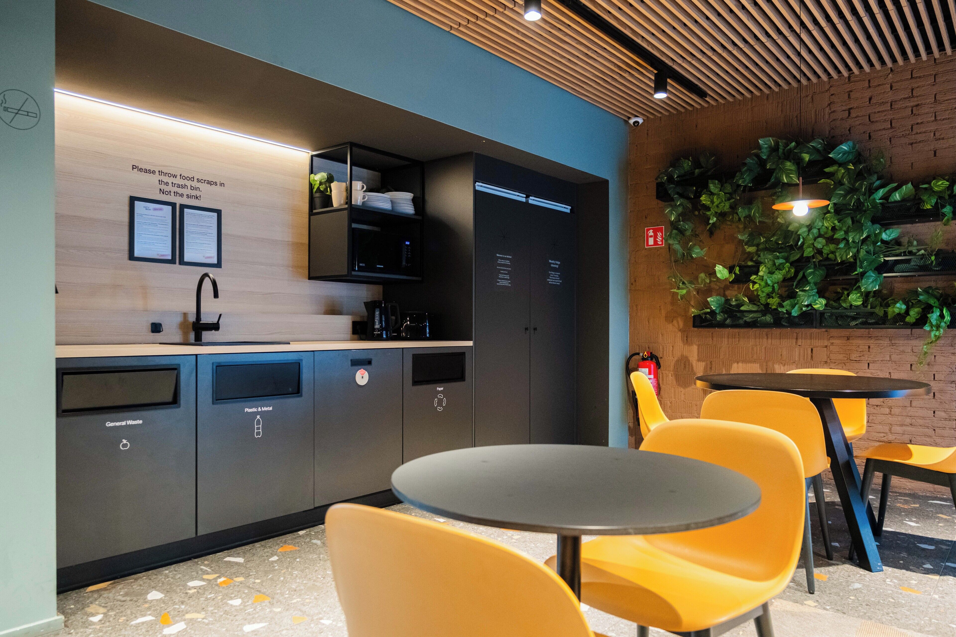 A modern kitchen area with a sink, microwave, and cabinets for storage. There are yellow chairs and a small table, and a wall-mounted plant decoration.