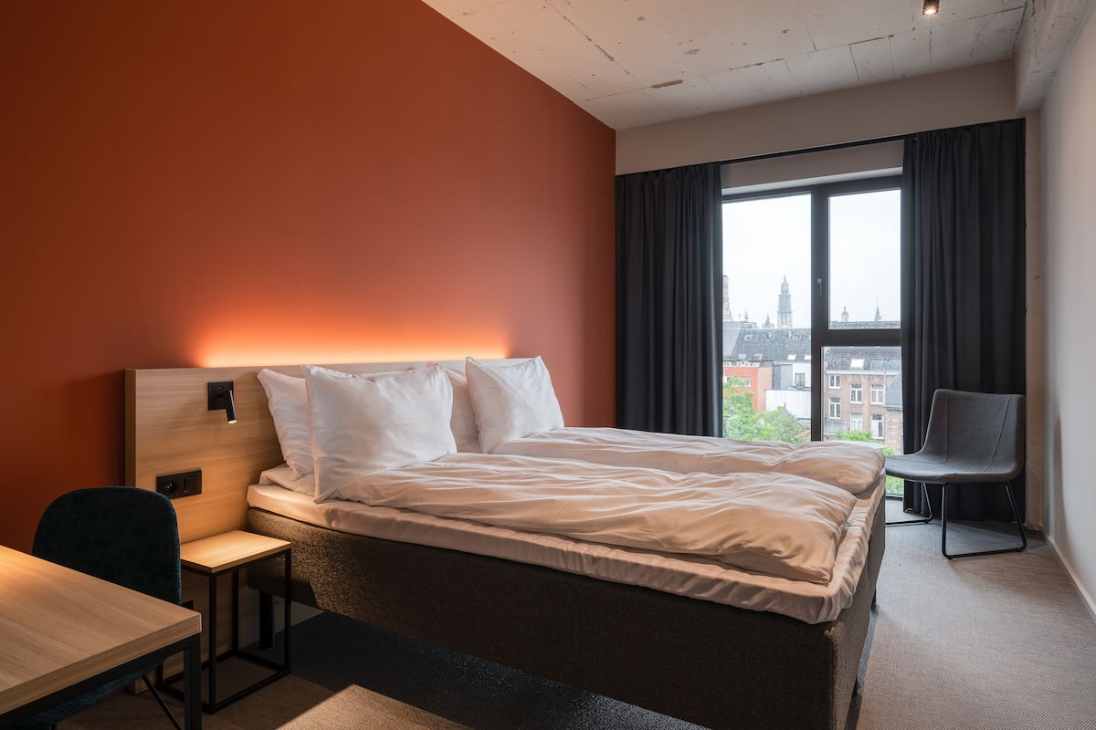 Een hotelkamer met een bed, een bureau, een stoel en een groot raam met uitzicht op de stad.