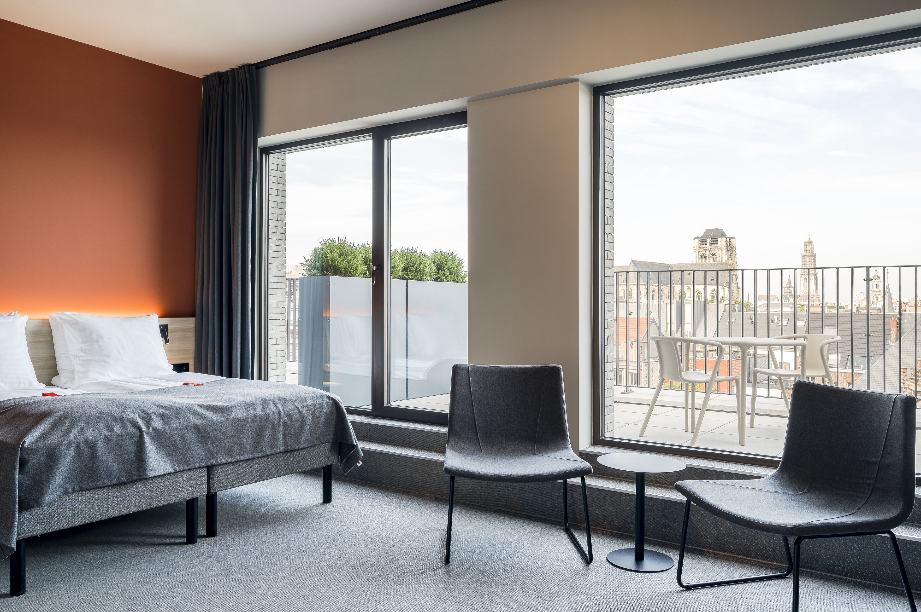 A hotel room with a bed, two chairs, a small table, and large windows offering a city view.