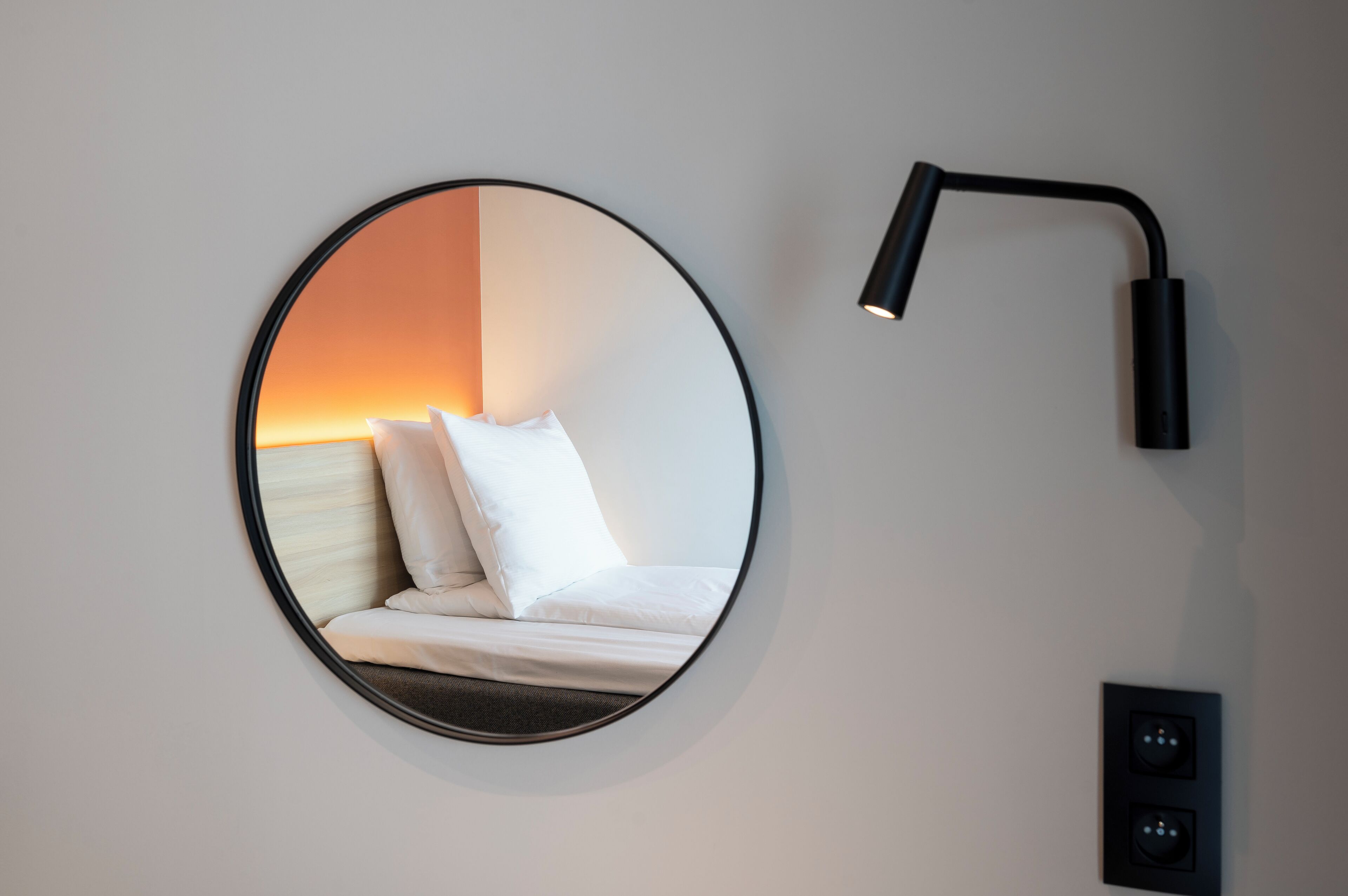 A circular mirror reflecting a bed with white bedding and a nightstand, a black wall-mounted lamp, and a power outlet.