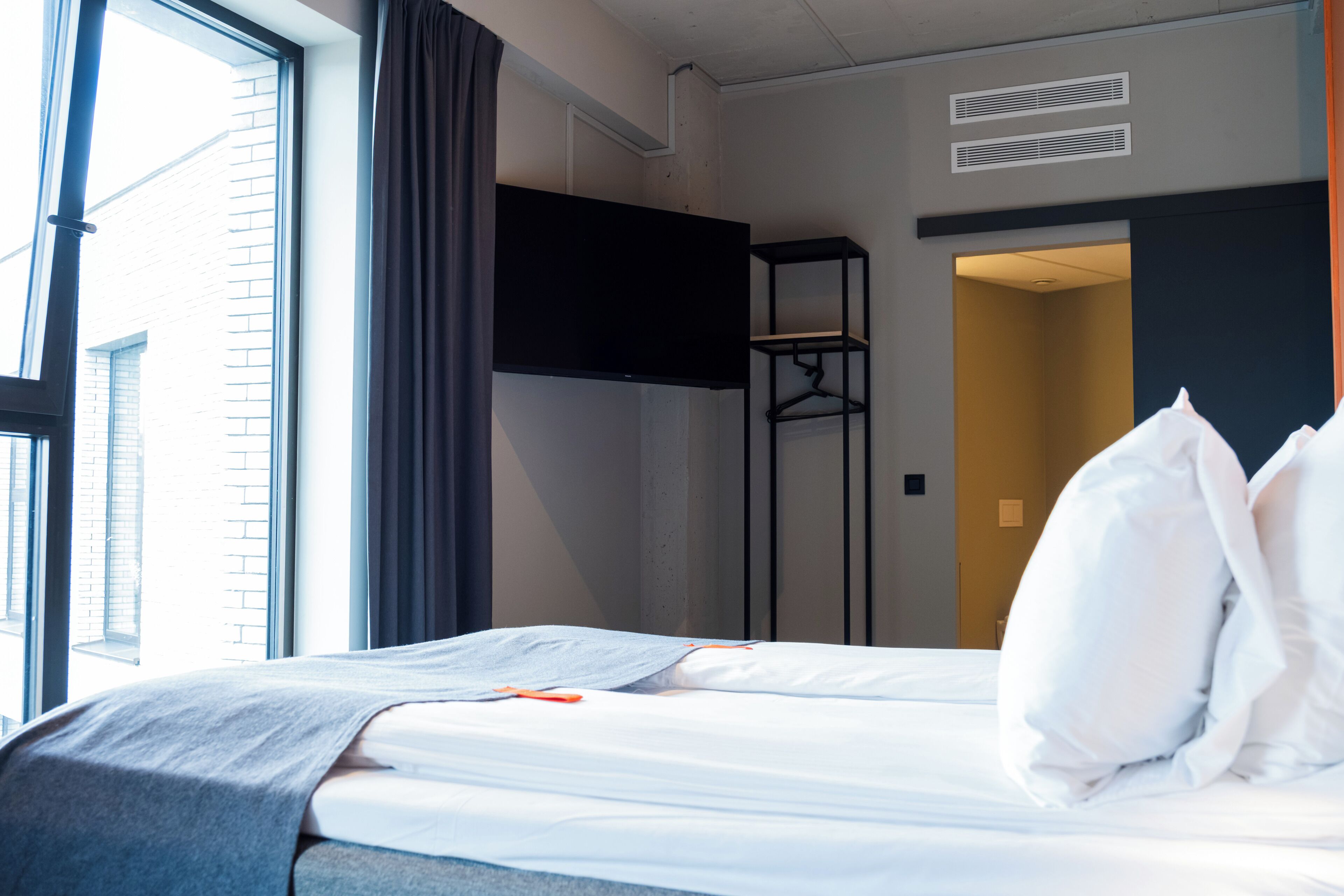 Une chambre d’hôtel avec un lit, un téléviseur fixé au mur, une fenêtre avec des rideaux et une porte donnant sur une autre pièce.