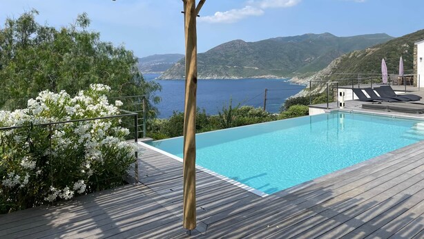 Une terrasse en bois avec une piscine, offrant une vue sur un paysage côtier avec des montagnes et un ciel dégagé.