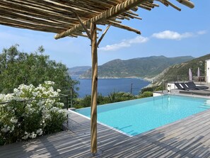 A wooden deck with a pool, overlooking a coastal landscape with mountains and a clear sky.
