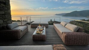 Une terrasse en bois, agrémentée de meubles en osier et d’une table dressée pour un repas, offrant une vue sur un paysage côtier.
