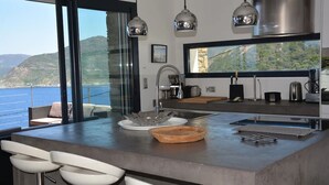 Une cuisine moderne dotée d’un grand îlot central, de tabourets de bar, de luminaires suspendus et offrant une vue sur la mer.
