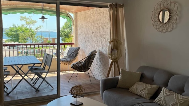 A modern living room with a grey sofa, a white coffee table, and a dining area with a table and chairs. A large window offers a view of the outdoors.