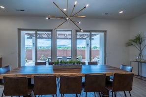 A spacious dining area with a large wooden table and six chairs, large windows offering a view of the outdoors, and a modern ceiling light fixture. - Arcadia 55 | Pool Table * PRIVATE Hot Tub * BBQ (Santa Clara)