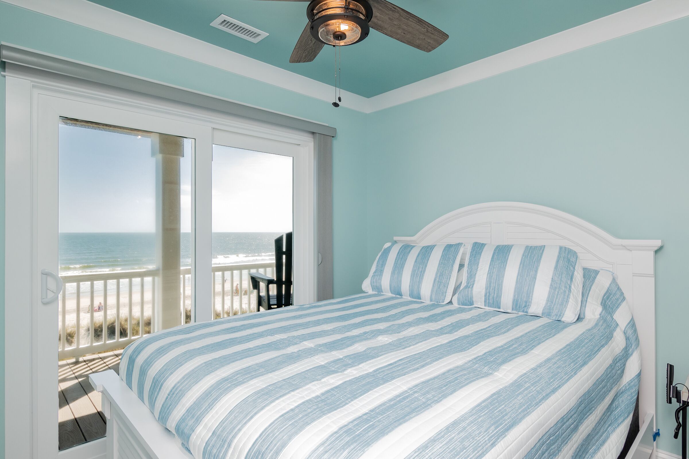 A bedroom with a bed, a ceiling fan, and a view of the beach through a sliding door.