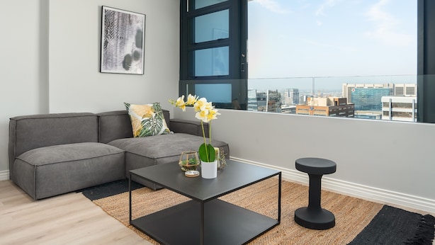Un salon moderne avec un canapé d’angle gris, une table basse noire et une petite table d’appoint. Un tableau abstrait encadré orne le mur, et une fenêtre offre une vue sur la ville.