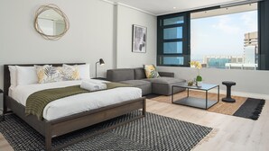 Une chambre moderne avec un lit, un canapé gris, une petite table et une grande fenêtre donnant sur la ville.